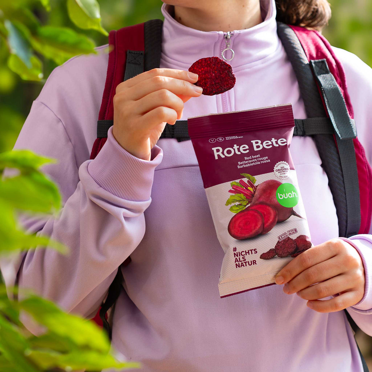 Wanderin in rosa Shirt hat eine Packung buah Rote Bete Chips in der Hand und isst einen Chip aus der Packung
