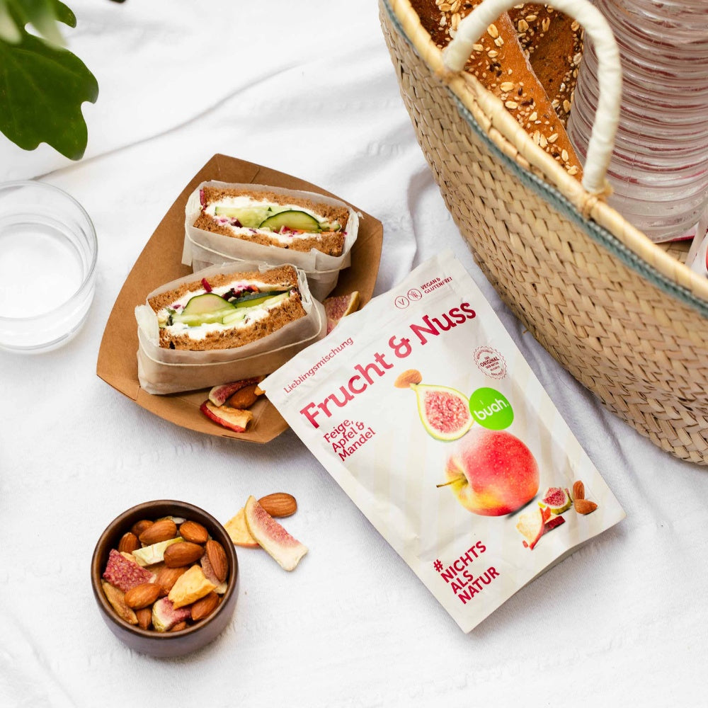 buah Frucht & Nuss Mischung Feige, Apfel und Mandel neben einem Picknickkorb und einem Sandwich