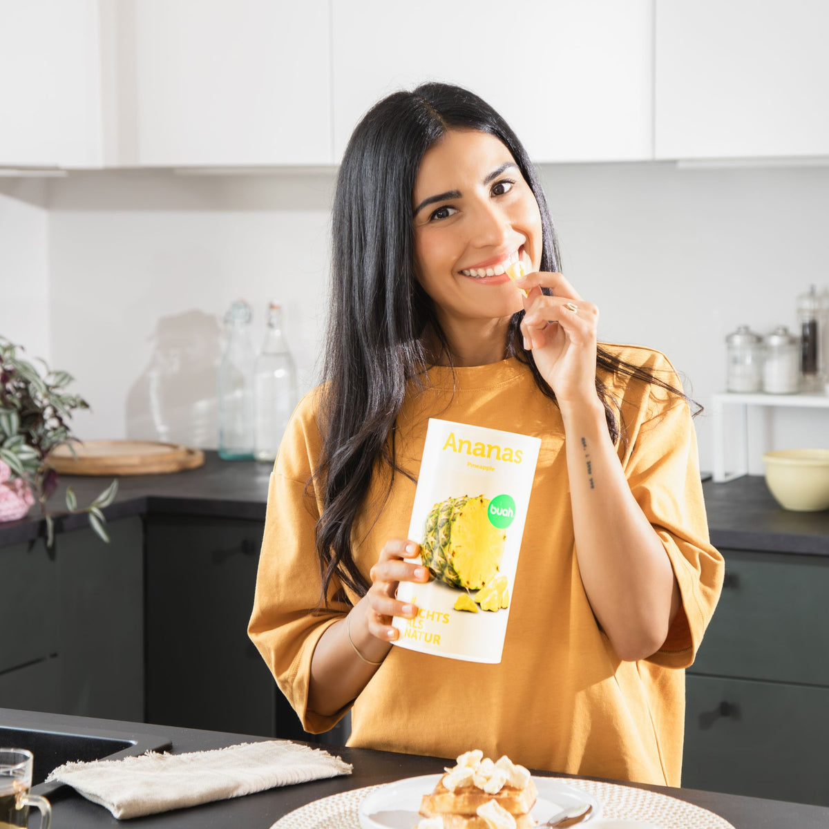 Junge Frau mit langen dunklen Haaren snackt ein Stueck gefriergetrocknete Ananas aus einer Dose buah Ananas und lacht in die Kamera