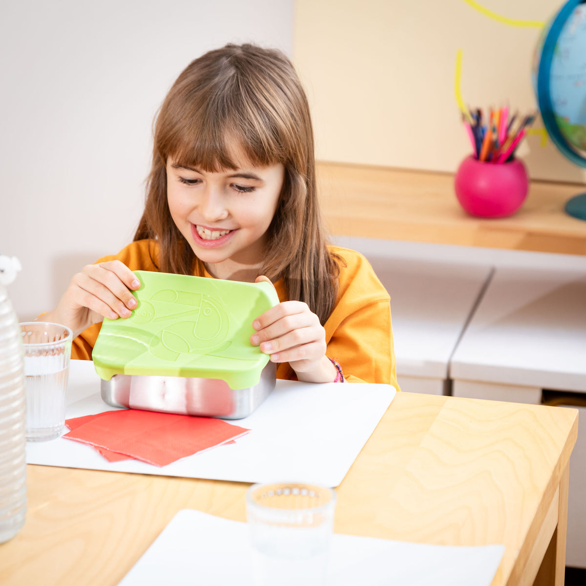 Junges Kind in der Grundschule sitzt am Tisch und oeffnet lachend die buah Lunchbox aus Edelstahl mit einem Silikon Deckel. 