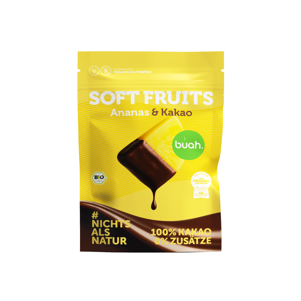 Ein gelber Beutel buah Soft Fruits der Sorte Ananas & Kakao steht vor einem gelben Hintergrund. Davor liegen einige softe, mit Schokolade überzogene Ananas-Stücke.