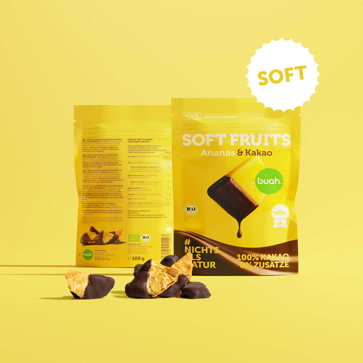 Ein gelber Beutel buah Soft Fruits der Sorte Ananas & Kakao steht vor einem gelben Hintergrund. Davor liegen einige softe, mit Schokolade überzogene Ananas-Stücke.