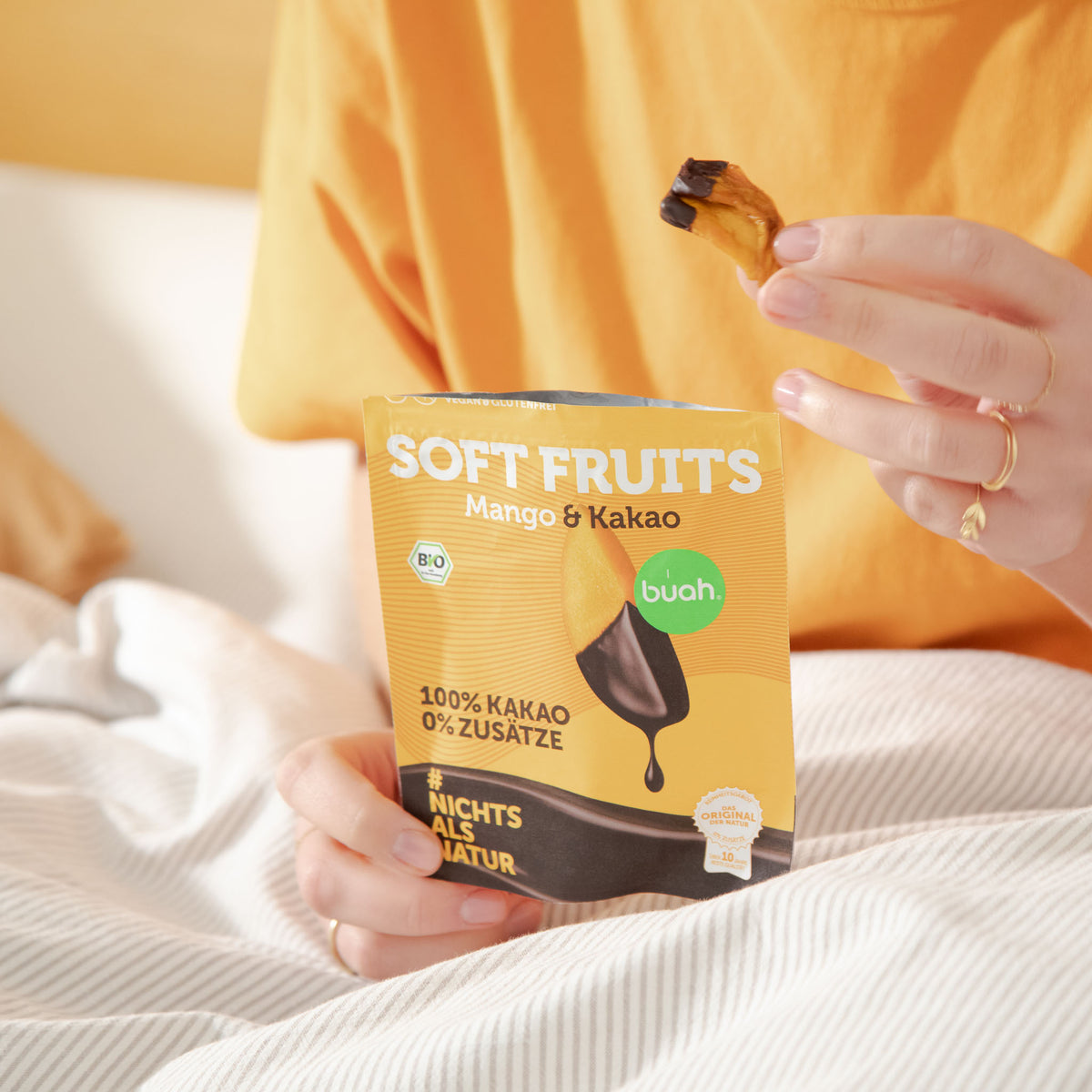 Nahaufnahme im Bett: Eine Person im orangefarbenen T-Shirt hält ein schokoliertes buah Soft Fruit Mango-Stück hoch. Vor ihr auf der Bettdecke steht der geöffnete Beutel der Sorte Mango & Kakao.