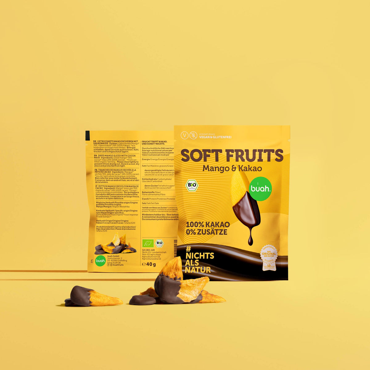 Zwei buah SOFT FRUITS Snackpacks Mango & Kakao vor gelbem Hintergrund. Davor liegen mehrere Mangoscheiben, teils mit dunkler Schokolade überzogen. Auf der Vorderseite steht „100% KAKAO 0% ZUSÄTZE“ und „#NICHTS ALS NATUR“.