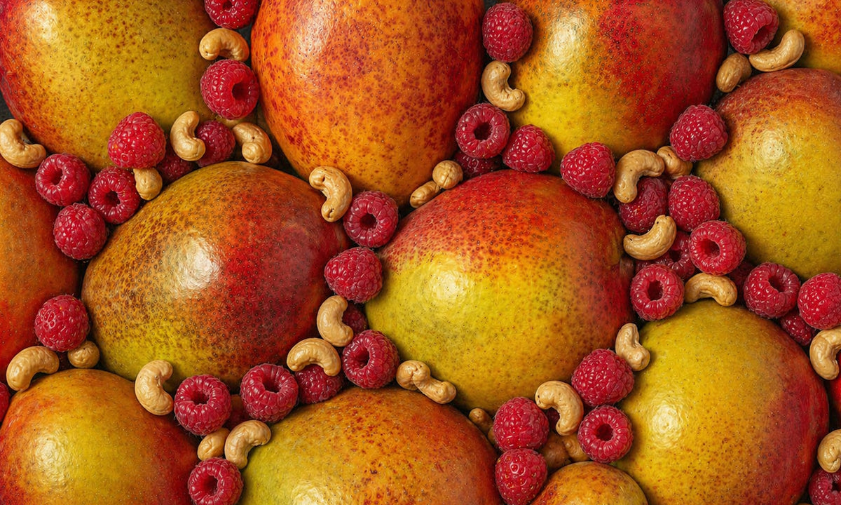 Eine farbenfrohe Nahaufnahme von vielen frischen, ganzen Mangos. Dazwischen liegen frische Himbeeren und helle Cashewkerne.