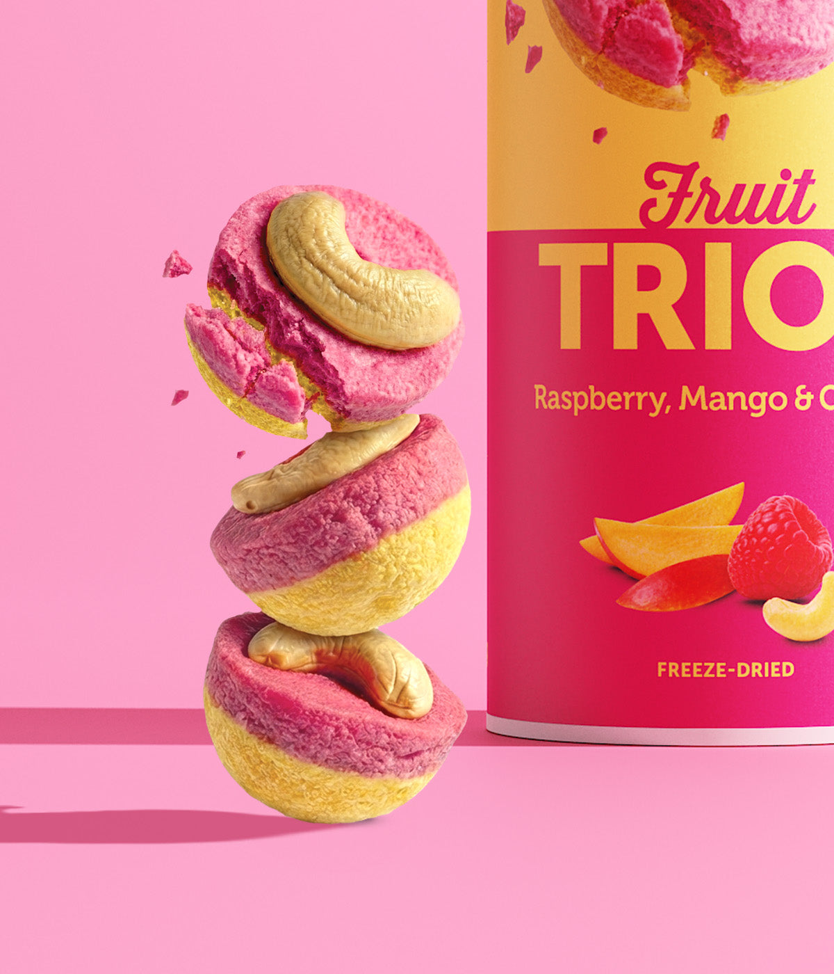 Ein Turm aus drei runden buah Fruit TRIOS Stücken der Sorte Raspberry, Mango & Cashew vor pinkem Hintergrund. Das oberste, pink-gelbe Fruchtstück zerbricht in der Luft. Auf jedem Stück liegt ein Cashewkern.